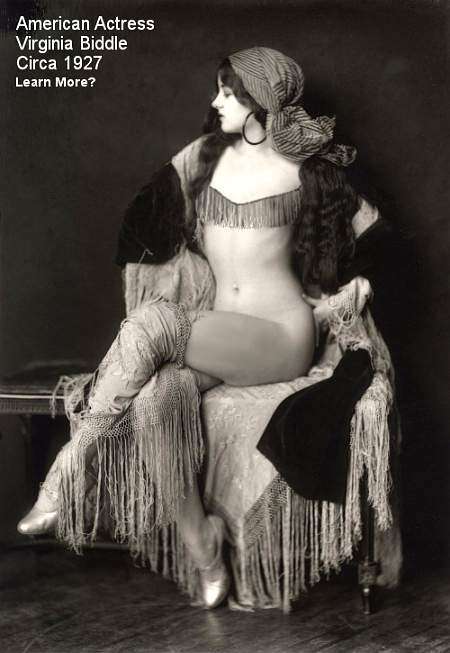 A black and white image of Virginia Biddle (1910 – 2003) scantily clad with what looks like Tarot Card - Psychic garb. She is seated on a couch and facing left in the frame. She has big hoop ear rings on, a head scarf and a shawl. Her legs are crossed, and she has a tassel garb across her chest. Most of her body is uncovered. She has very long dark hair and she is a very attractive woman.